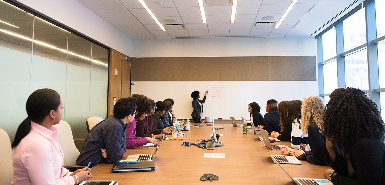 Team of people in a meeting looking at a woman writing on a whiteboard 